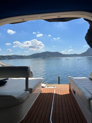 Une terrasse de bateau avec des sièges et une vue sur un lac paisible, avec des bateaux et des collines à l’horizon.