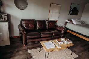 A room with a brown leather sofa, a wooden coffee table with books and decorative items, a bed with patterned bedding, a white refrigerator, and a wall-mounted mirror. - The Aspen Abode - Cozy 1-bedroom cabin in peaceful Kalispell with AC (Kalispell)