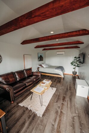 A room with a wooden ceiling, a brown leather sofa, a bed with a white headboard, a small table with a plant, and a white cabinet. - The Aspen Abode - Cozy 1-bedroom cabin in peaceful Kalispell with AC (Kalispell)