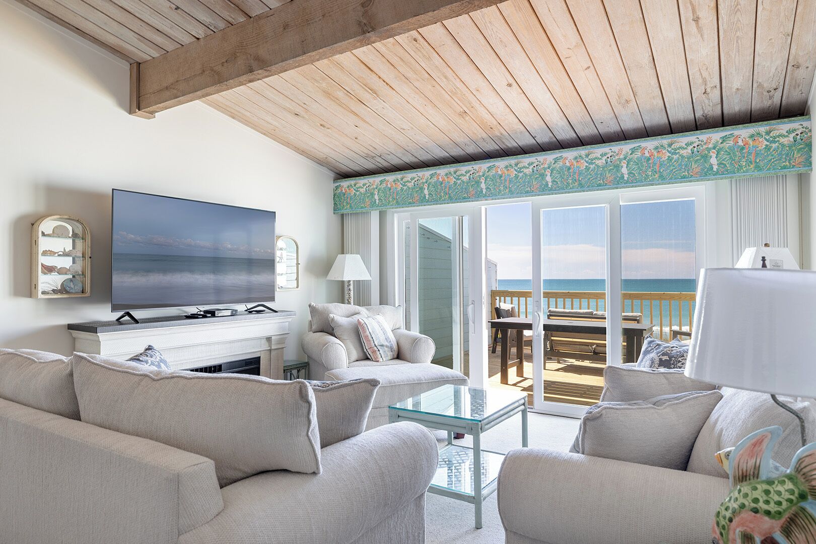 A living room with a television, a fireplace, a sofa, a glass coffee table, and a view of the beach through large sliding glass doors.