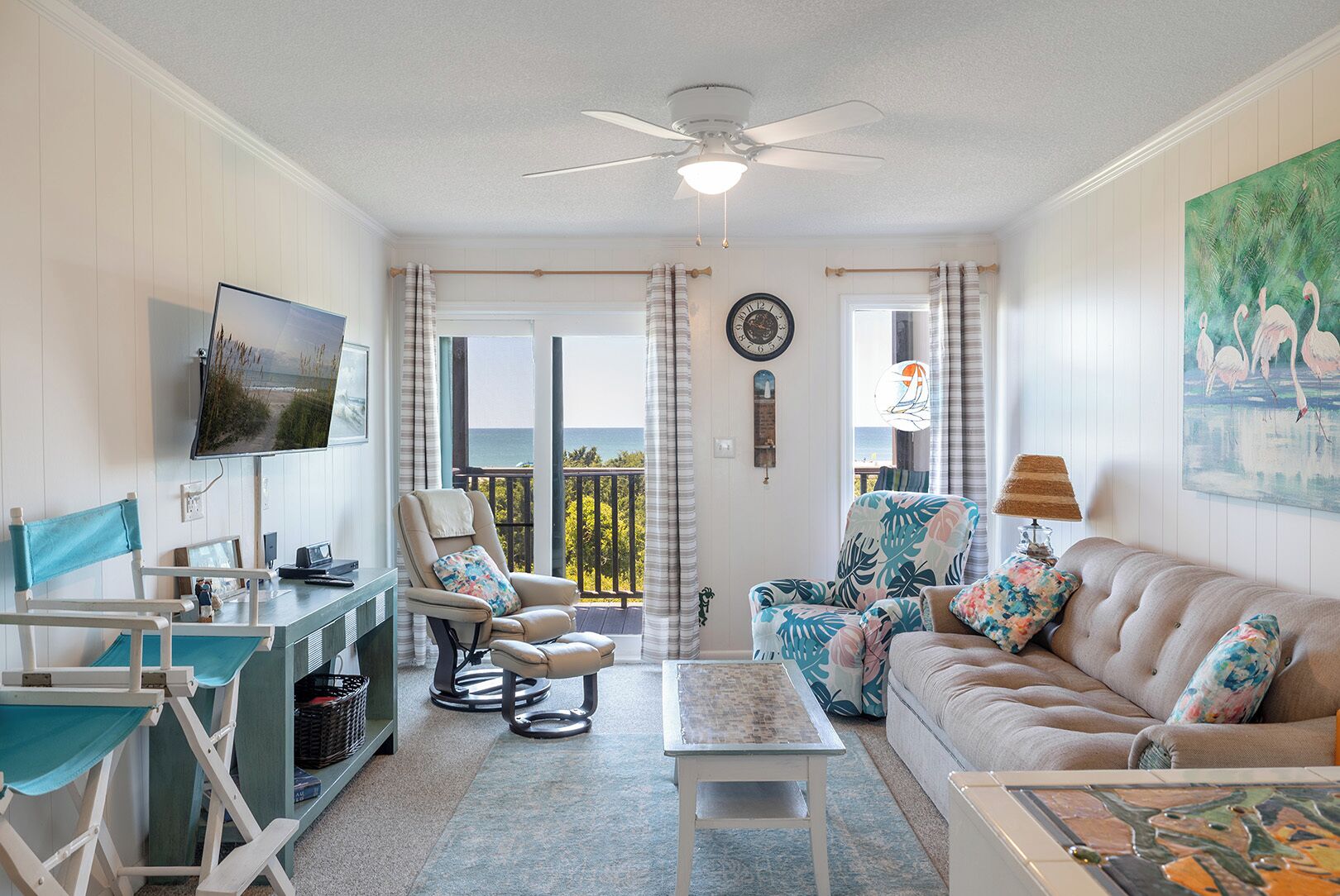 A living room with a TV, a sofa, a chair, a table, and a balcony with a view.