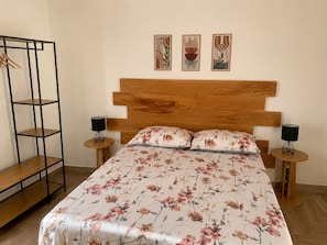 A bedroom with a wooden headboard, floral bedding, a wooden side table, a hanging rack, and three framed artworks on the wall.
