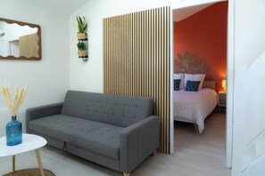 Une chambre moderne avec un canapé gris, une petite table ronde ornée d’un vase bleu, un lit avec une literie blanche et des coussins décoratifs, et un panneau en bois vertical.