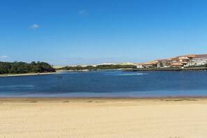 Une plage côtière avec une jetée qui s’avance dans l’eau, une rive sablonneuse et, au loin, un bâtiment surmonté d’un toit rouge.