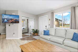 A modern living room with a flat-screen TV, a wooden coffee table, a white sofa with blue cushions, and a large window with curtains. - Group Getaway in Windsor | Sleeps 20 (Windsor)