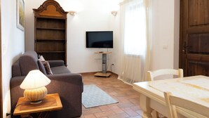 A living room with a sofa, a table, a lamp, a TV, and a bookshelf. - Nice apartment for 4 people with WIFI, pool, A/C, TV, patio and pets allowed (Panicale)