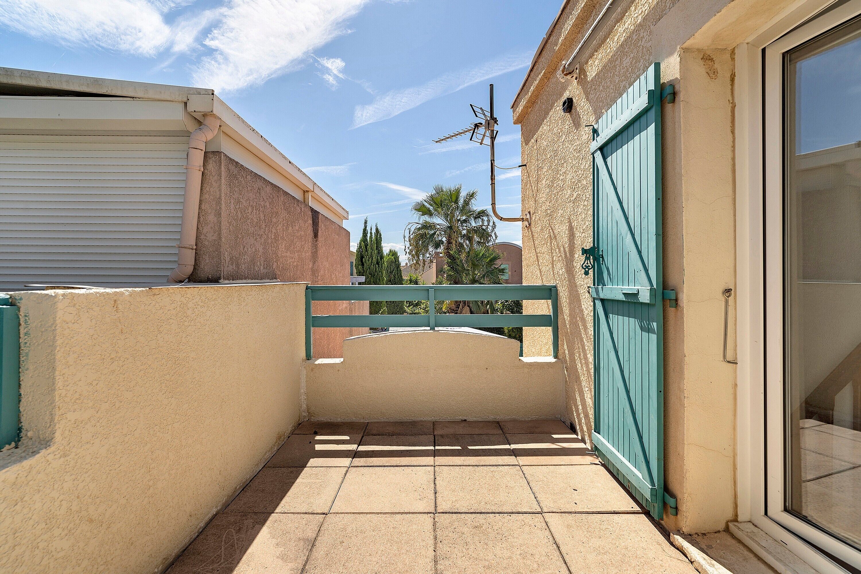 A narrow outdoor corridor with a tiled floor, a metal gate, and a building with a beige wall and a green door.