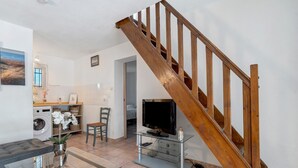 Una sala de estar con un sofá, una mesa de centro de vidrio, un televisor sobre un mueble bajo, una escalera de madera y una lavadora.