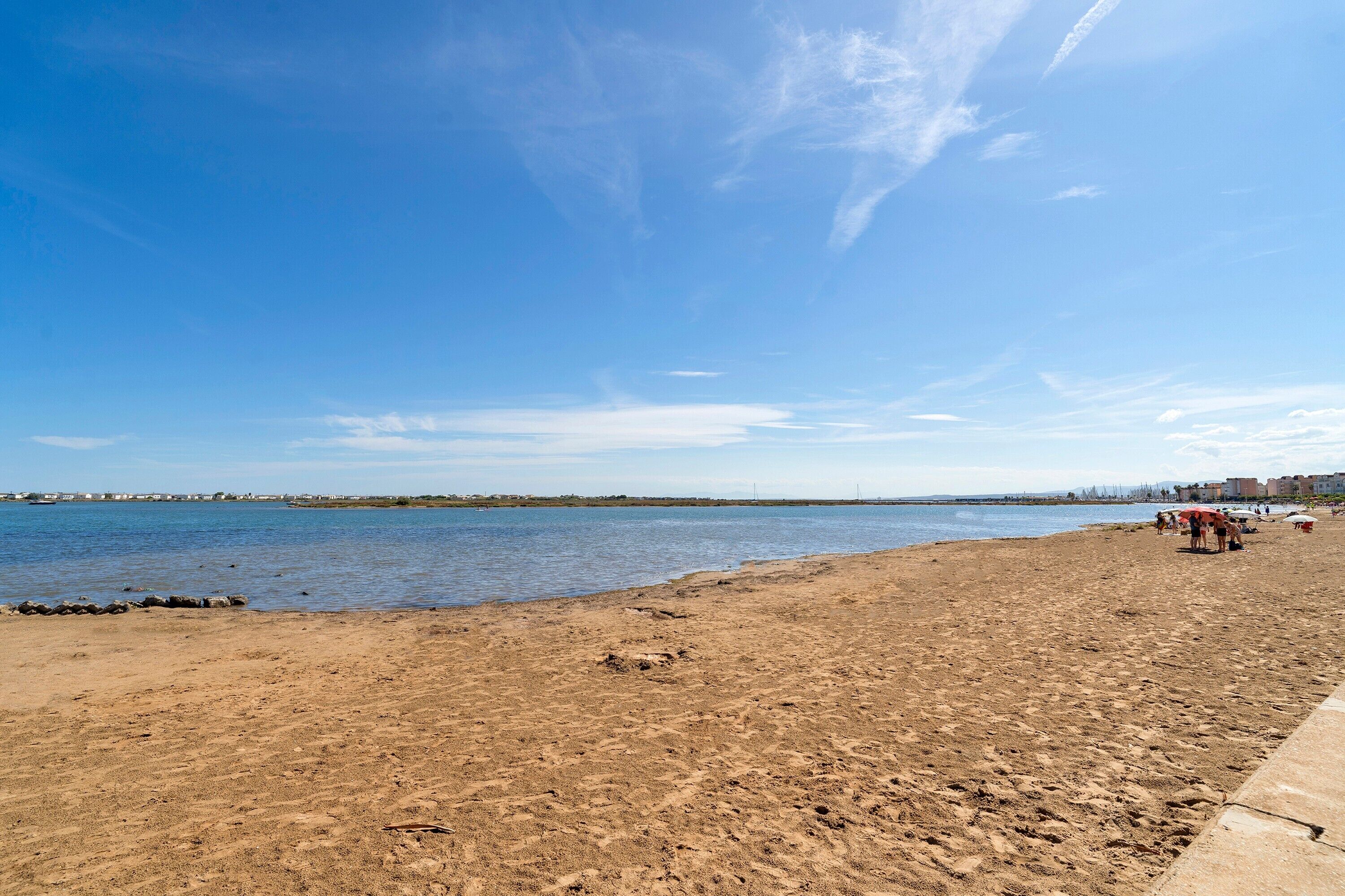 Une plage avec un ciel dégagé, une eau calme et une côte lointaine parsemée de bâtiments.