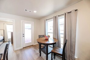 A room with a wooden dining table, chairs, a white door, and a window with curtains. - The Jade - Cozy Lubbock Getaway - Sleeps 6 (Lubbock)