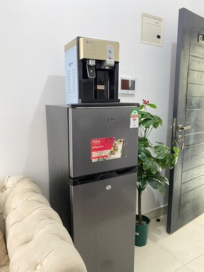 Ein kompakter Kühlschrank mit Wasserspender und einem Aufkleber von Von, neben einem Blumentopf in einem Raum mit einer beigen Couch und einer Tür.
