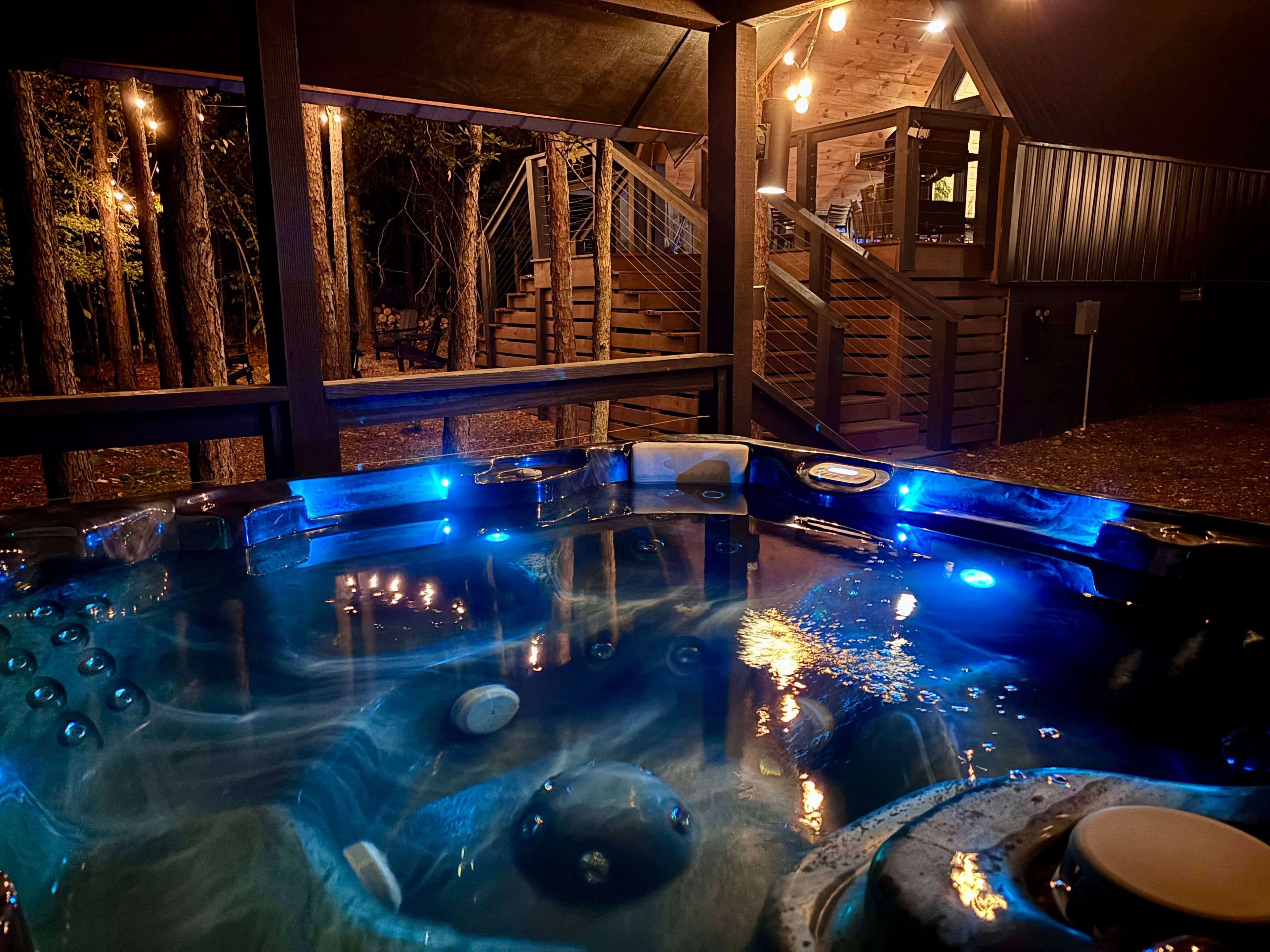 Ein Whirlpool mit blauer Beleuchtung, umgeben von einer Holzdielen-Terrasse und Treppen, die zu einem Gebäude führen.