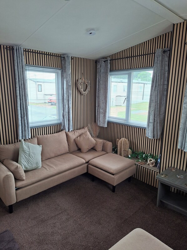 Ein Wohnzimmer mit einem beigefarbenen Ecksofa, zwei Fenstern mit Jalousien, gestreifter Tapete und einer Deckenleuchte.
