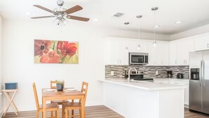 A modern kitchen with white cabinetry, a central island, a ceiling fan, and a dining area with wooden chairs and a table. - Reed Unit B - Home & Private Rooms (Houston)