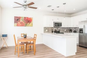 Eine moderne Küche mit weißen Schränken, einer zentralen Kochinsel, einem Deckenventilator und einem Essbereich mit Holztischen und -stühlen.