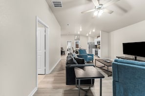 A modern living room with a sofa, coffee table, and TV. There is a kitchen in the background with a microwave and cabinets.