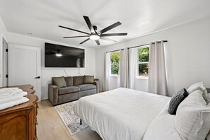 A bedroom with a bed, a ceiling fan, a TV mounted on the wall, a sofa, and a window with curtains. - Seaside Ridge Above the Fog | Hot Tub, Ocean View (Gualala)