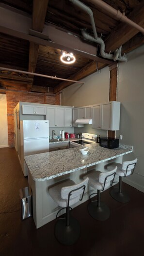 A kitchen with a granite countertop, white cabinets, a stainless steel refrigerator, a stove, and a sink. There are three bar stools and a ceiling light fixture. - Freshly renovated Studio Loft in the heart of Corktown. (Detroit)
