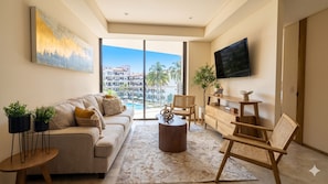 A modern living room with a sofa, coffee table, and chairs, a flat-screen TV, and a balcony with a pool view.