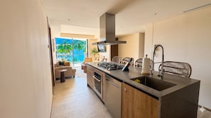 Uma cozinha moderna com uma ilha central, forno embutido e uma janela grande com vista para uma varanda, palmeiras e o mar.
