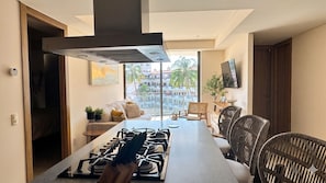 Une cuisine moderne équipée d’une cuisinière à gaz, d’une grande fenêtre offrant une vue sur les immeubles et les palmiers, et d’un coin repas avec des chaises en osier.