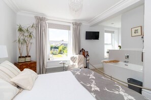A hotel room with a bed, a radiator, a TV, a bathroom with a bathtub, and a window with curtains.