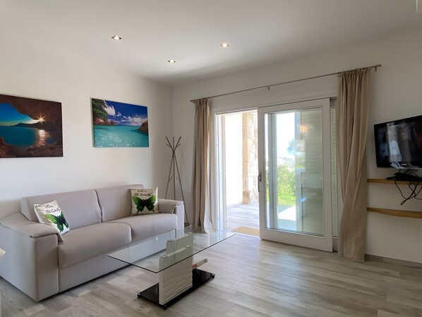Een moderne woonkamer met een bank, een glazen salontafel, een flatscreen televisie en een schuifpui naar een buitenruimte.