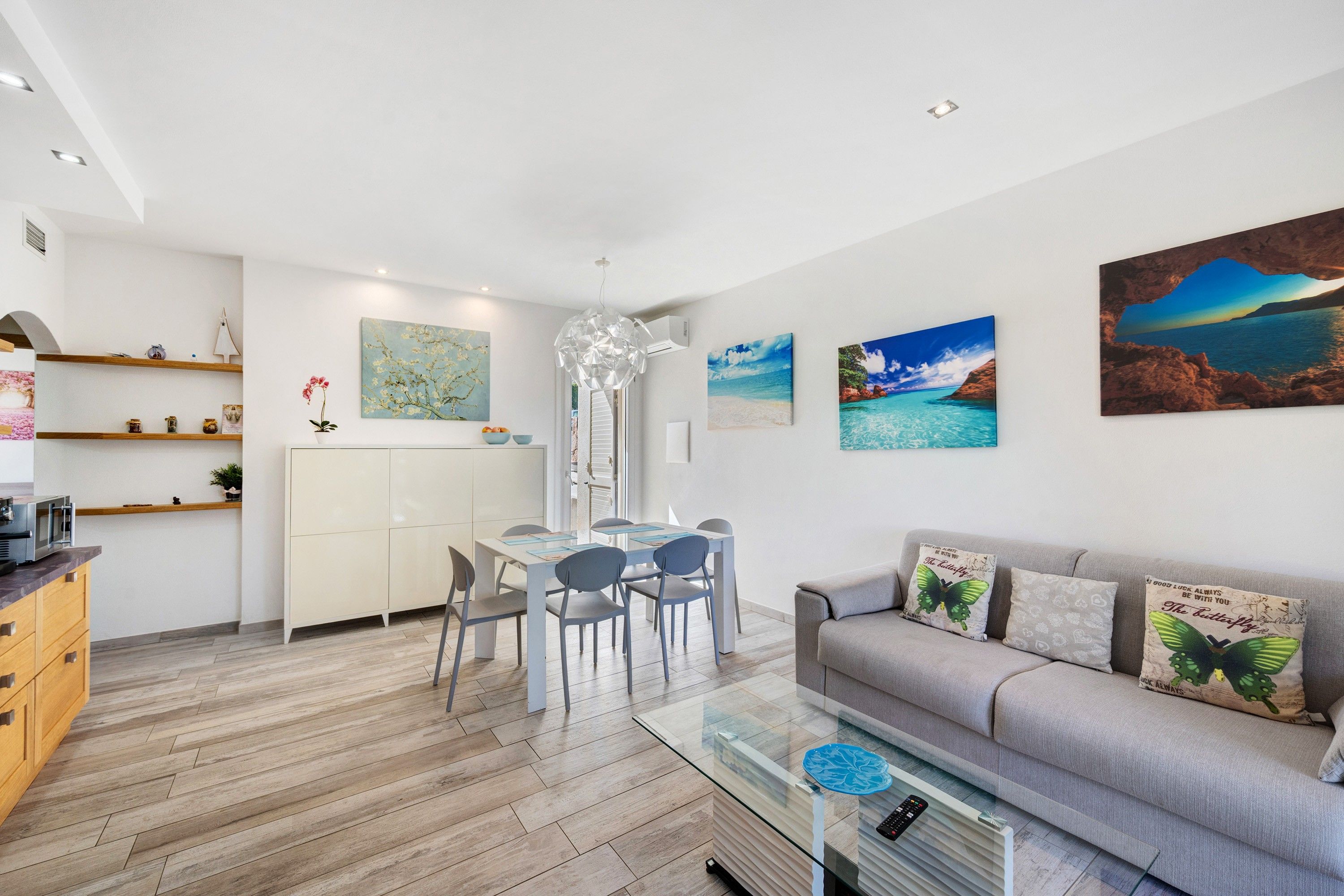 Ein modernes Wohnzimmer mit einem Glastisch, einem grauen Sofa mit Zierkissen und einem Essbereich mit weißem Tisch und Stühlen.