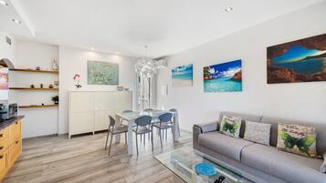Ein modernes Wohnzimmer mit einem Glastisch, einem grauen Sofa mit Zierkissen und einem Essbereich mit weißem Tisch und Stühlen.