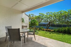 Un patio exterior con una mesa redonda y sillas, una pared blanca con textura, y vistas a una piscina y follaje.