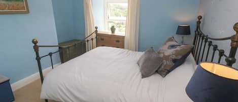 A bedroom with a bed, a nightstand, a lamp, a window with curtains, and a painting on the wall.