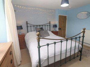 A bedroom with a metal bed frame, a wooden door, a bedside table with a lamp, and string lights on the wall. - Greenways (Perranporth)