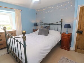 A bedroom with a bed, two bedside tables with lamps, a dresser, a window with curtains, and a door. - Greenways (Perranporth)
