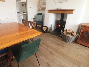 A dining area with a wooden table and chairs, a fireplace with logs, and a kitchen in the background. - Greenways (Perranporth)