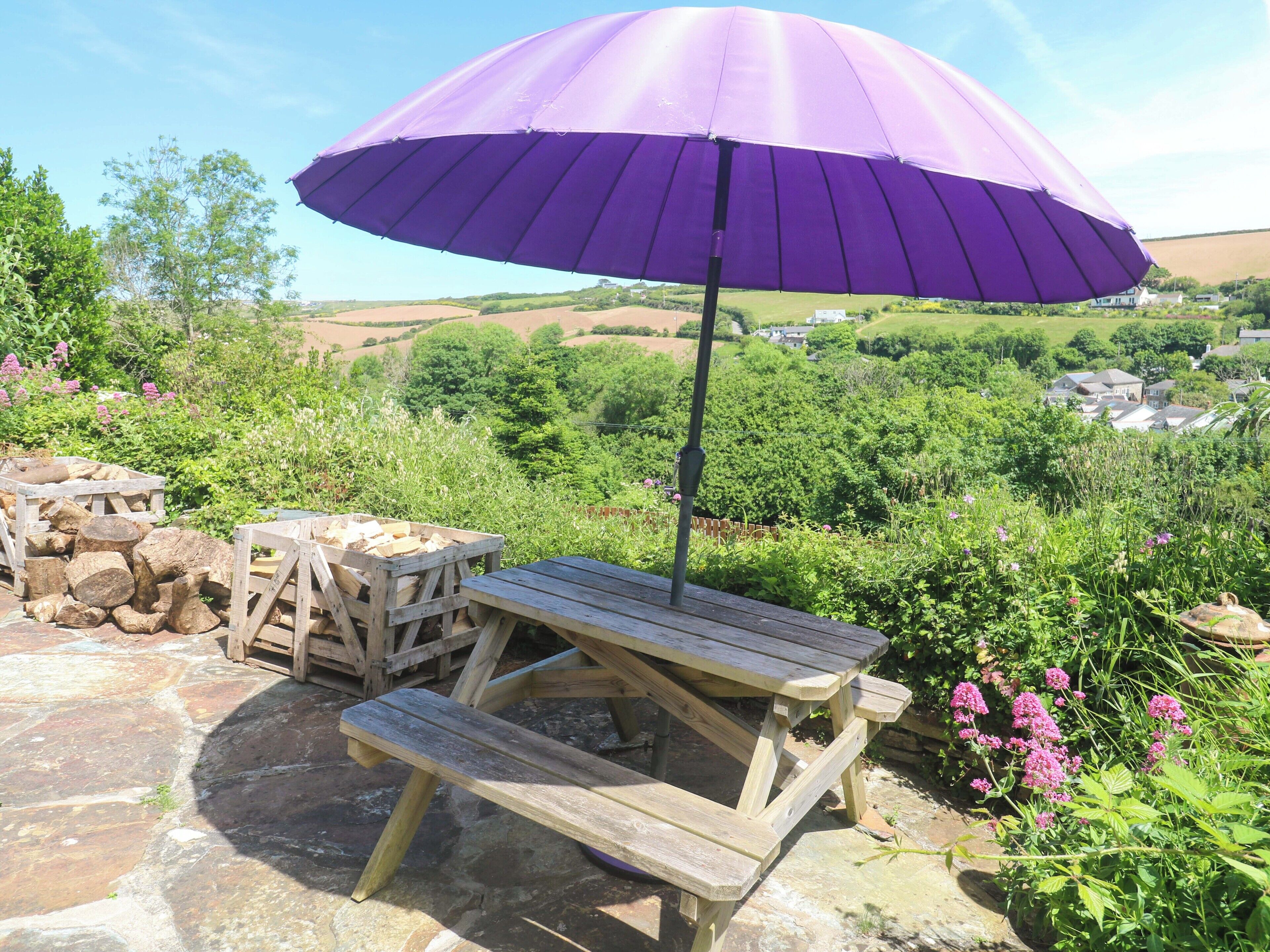Un conjunto de mesa y bancos de madera al aire libre, con un paraguas morado, rodeado de follaje y una vista panorámica.