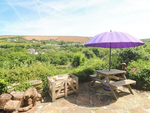 A wooden table and bench set under a purple umbrella, surrounded by greenery and stone flooring. - Greenways (Perranporth)