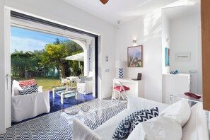A modern hotel room with a large sliding glass door leading to a garden, a bed with patterned bedding, a desk with a laptop, and a small seating area. - De Vivo Realty - Blue Skyline Villa in Sorrento Peninsula (Marciano)