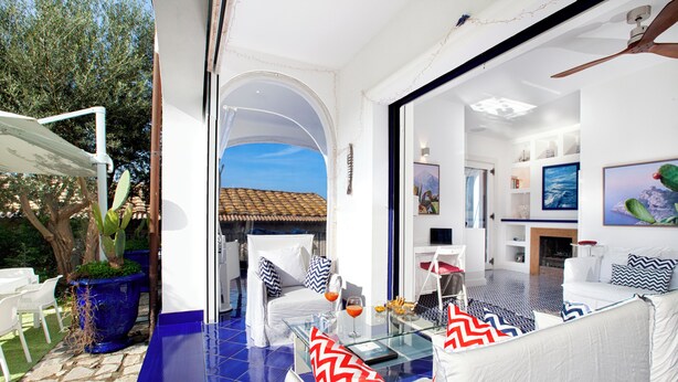 A modern living room with a glass coffee table, white sofas with patterned cushions, and a view of the outdoors. - De Vivo Realty - Blue Skyline Villa in Sorrento Peninsula (Marciano)