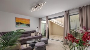Een moderne woonkamer met een bank, een voetenbankje en een salontafel, een schilderij aan de muur en een trap naar een balkon.