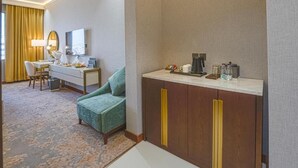 A hotel room with a green armchair, a desk with a lamp, a coffee maker, and a minibar. - Grand Iva Hotel Abha (Abha)