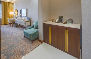 Habitación de hotel con un sillón verde, un escritorio con lámpara, cafetera y minibar.
