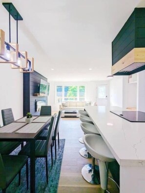 Une cuisine moderne avec un plan de travail blanc, un coin repas comprenant une table et des chaises, et une vue sur l’extérieur.