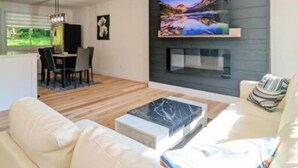 Uma sala de estar moderna com sofá, mesa de centro, TV fixada na parede e área de jantar com mesa e cadeiras.