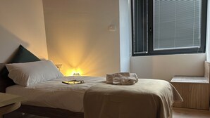 Una cama bien hecha con edredón blanco y una toalla doblada, una mesita de noche con lámpara, y una ventana con persianas.