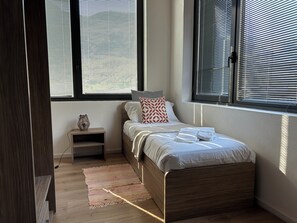 Ein Schlafzimmer mit Bett, Nachttisch, Lampe und Fenstern mit Jalousien.