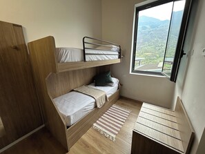 Ein kleines Zimmer mit Etagenbett, einem Schreibtisch, einem Fenster mit Blick auf eine gebirgige Landschaft und einem Holzschrank.