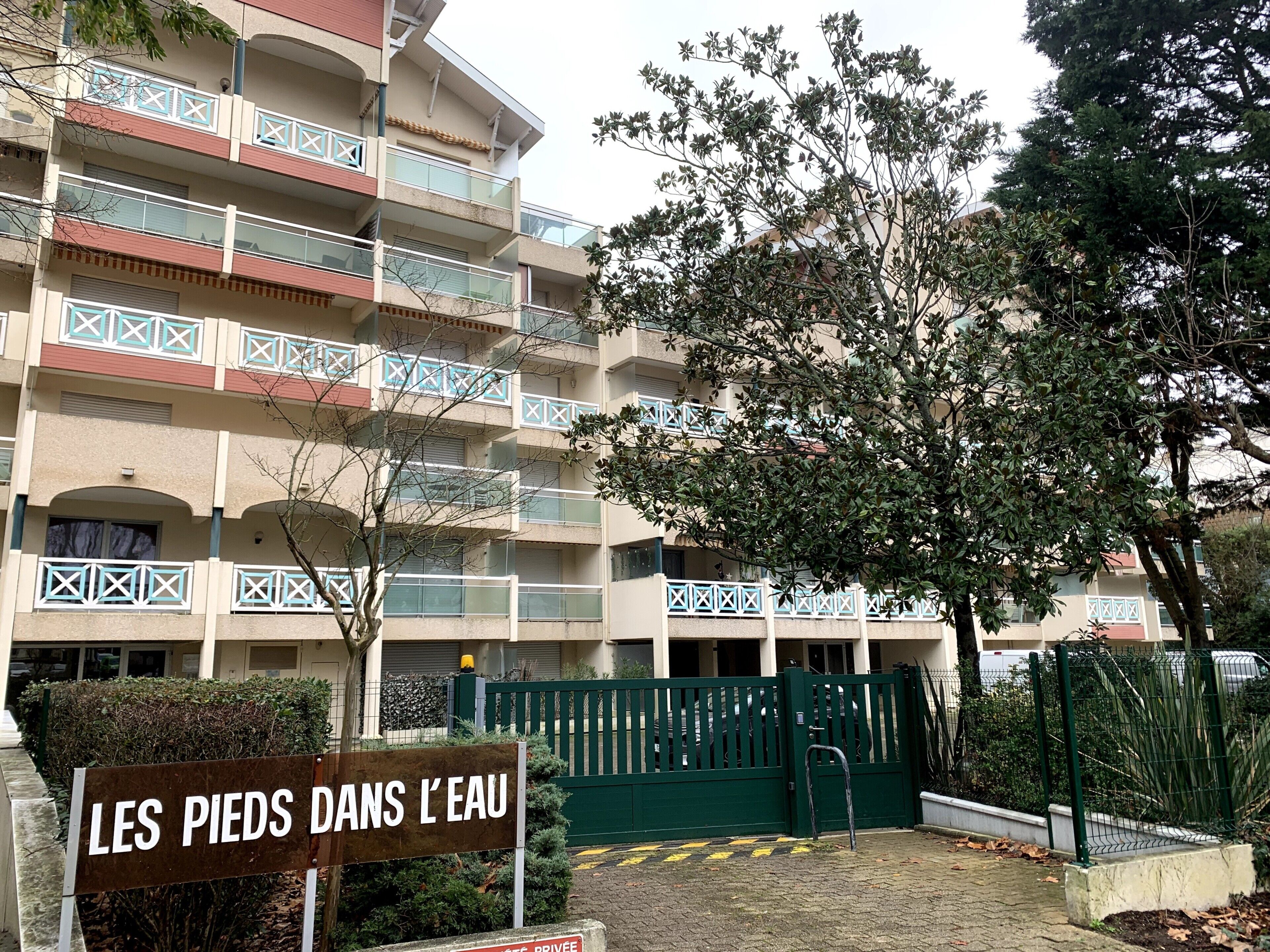 A multi-story building with balconies, a sign reading 'LES PIEDS DANS L'EAU,' and a gated entrance.