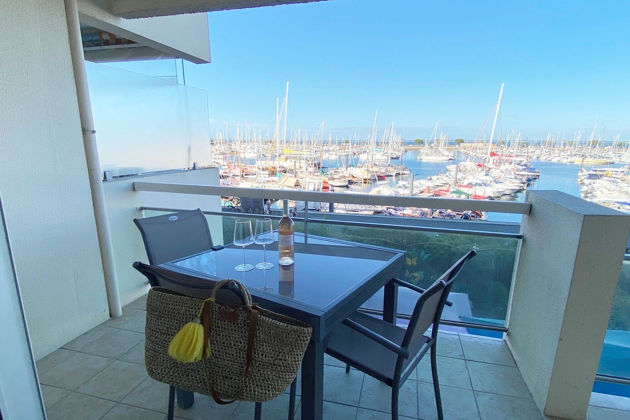 A balcony with a table and chairs, a view of a marina with boats, and a yellow bag hanging on the chair.