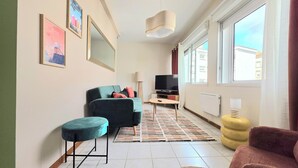 Un salon moderne avec un canapé, un pouf, un téléviseur et une grande fenêtre.