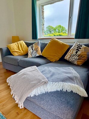 Ein graues Sofa mit gemusterten Kissen, einer weißen, fransigen Decke und Blick auf Grün durch ein Fenster.